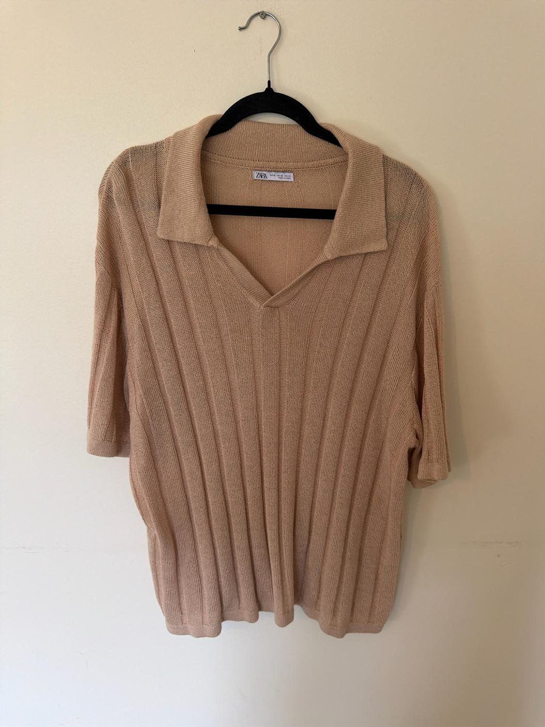 Beige short-sleeved knitted polo shirt with a ribbed texture, hanging on a black hanger against a light background. ZARA is written on the label.