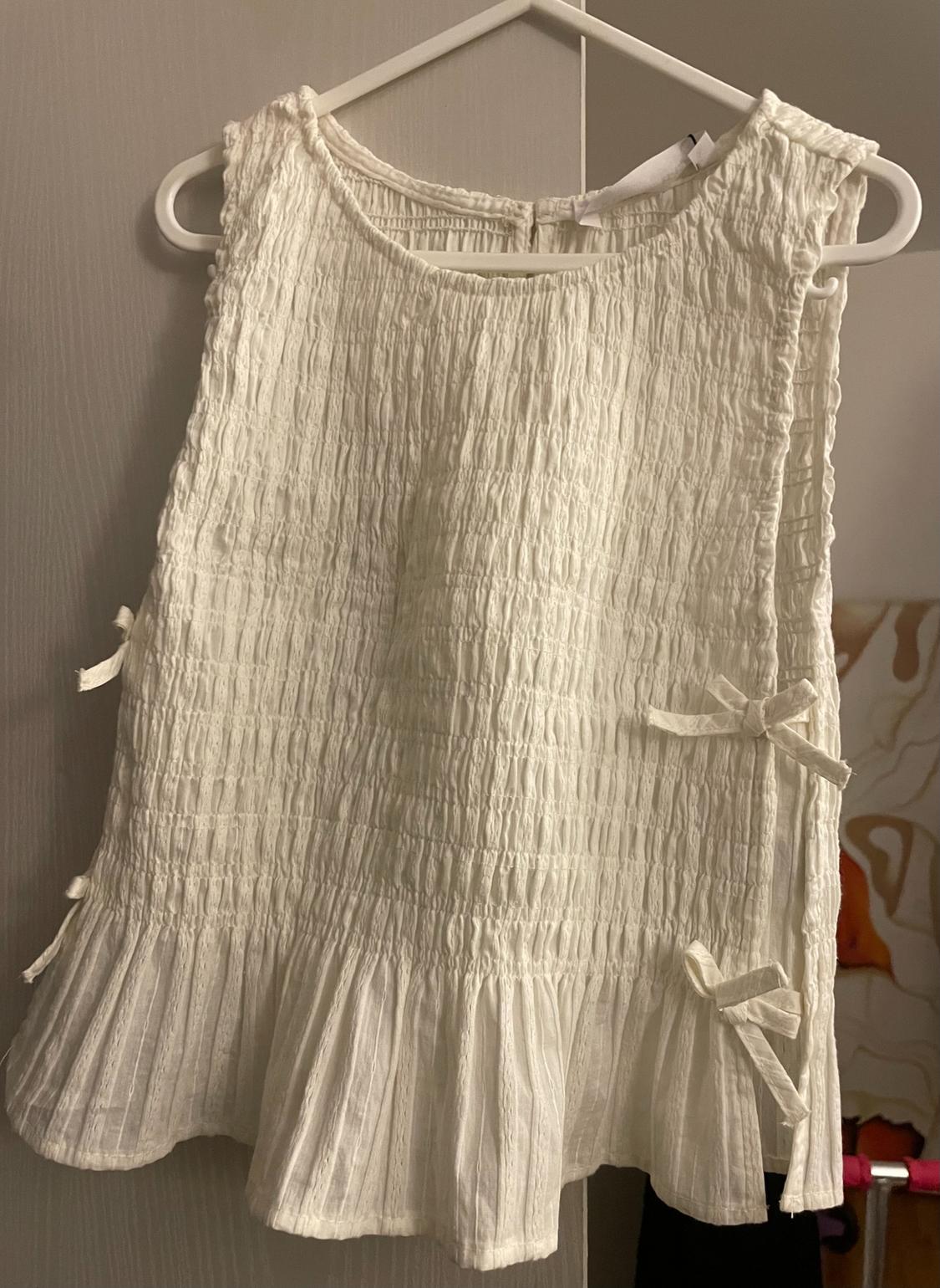 White crinkled fabric blouse with ruffles on the hem and decorative bows on the side, hanging on a white hanger.