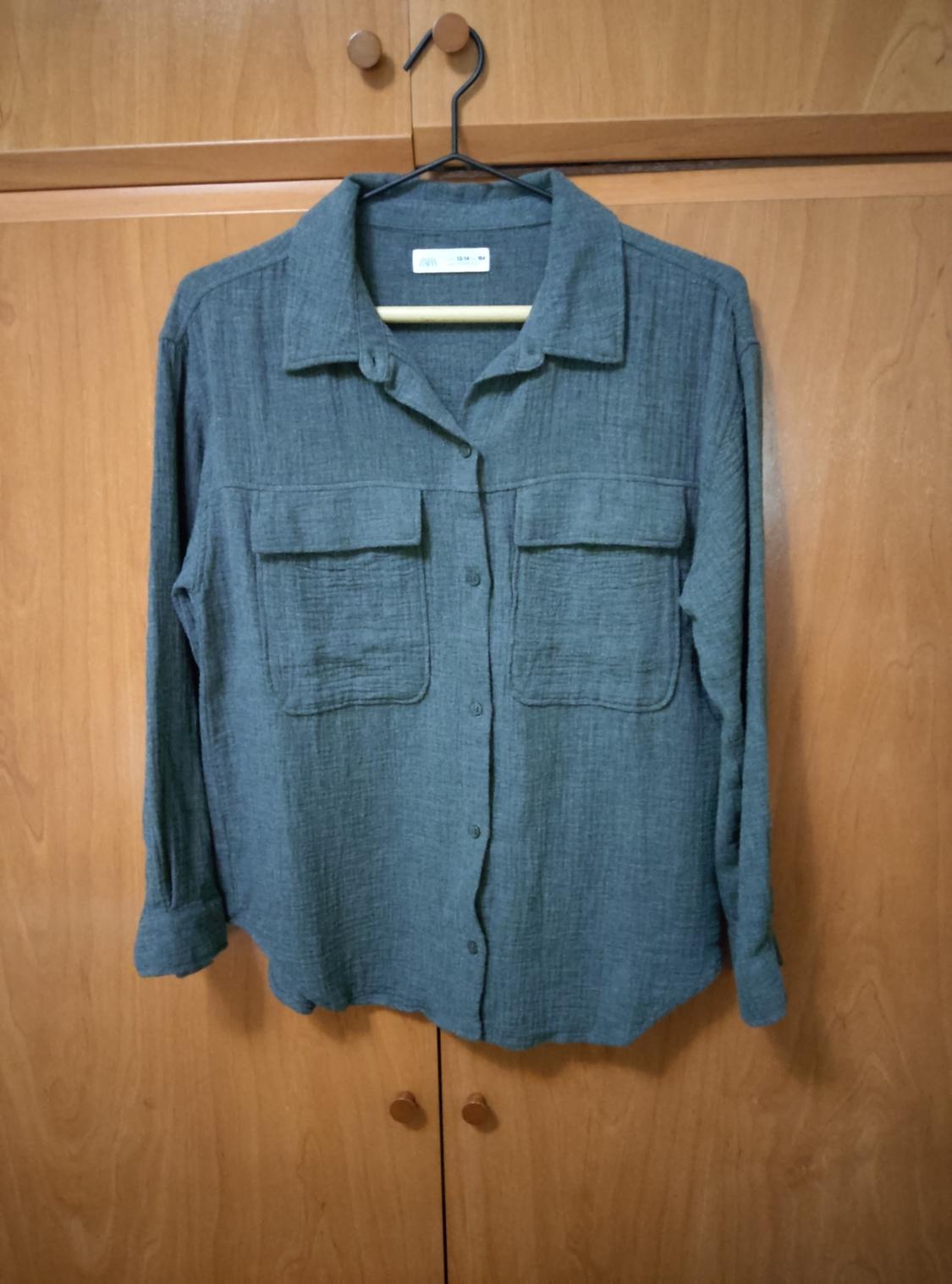 Long-sleeved grey shirt with front pockets, hanging on a hanger in front of a wooden wardrobe.