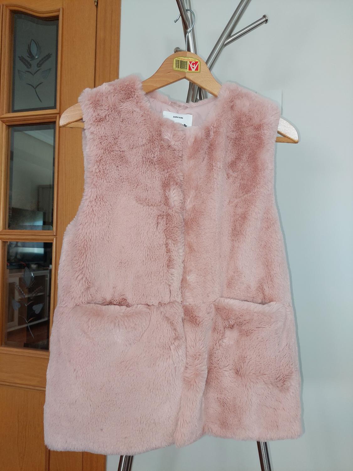 Light pink plush sleeveless gilet hanging on a wooden hanger, with a wooden door and white wall in the background.