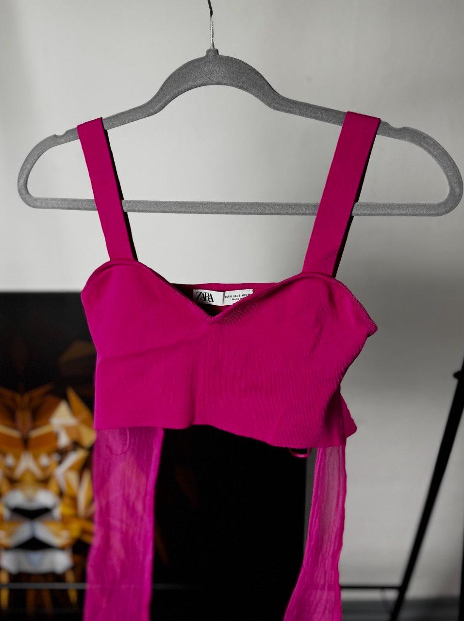 Fuchsia pink cropped top with a sweetheart neckline, wide straps and sheer fabric at the hem, on a grey hanger. ZARA label. Blurred background.