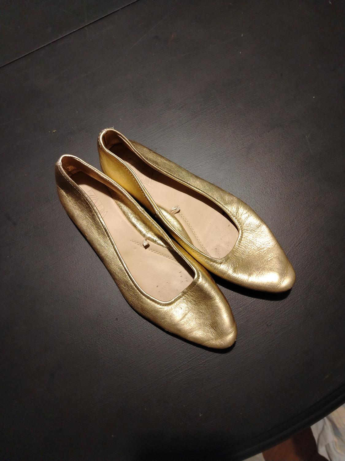 Gold flat shoes on a dark surface, viewed from above.