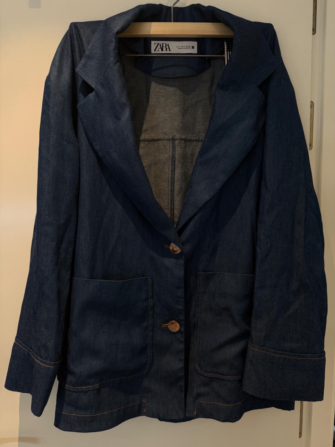 Blue denim jacket hanging on a wooden hanger, with two large pockets and brown buttons. Visible label: Zara.