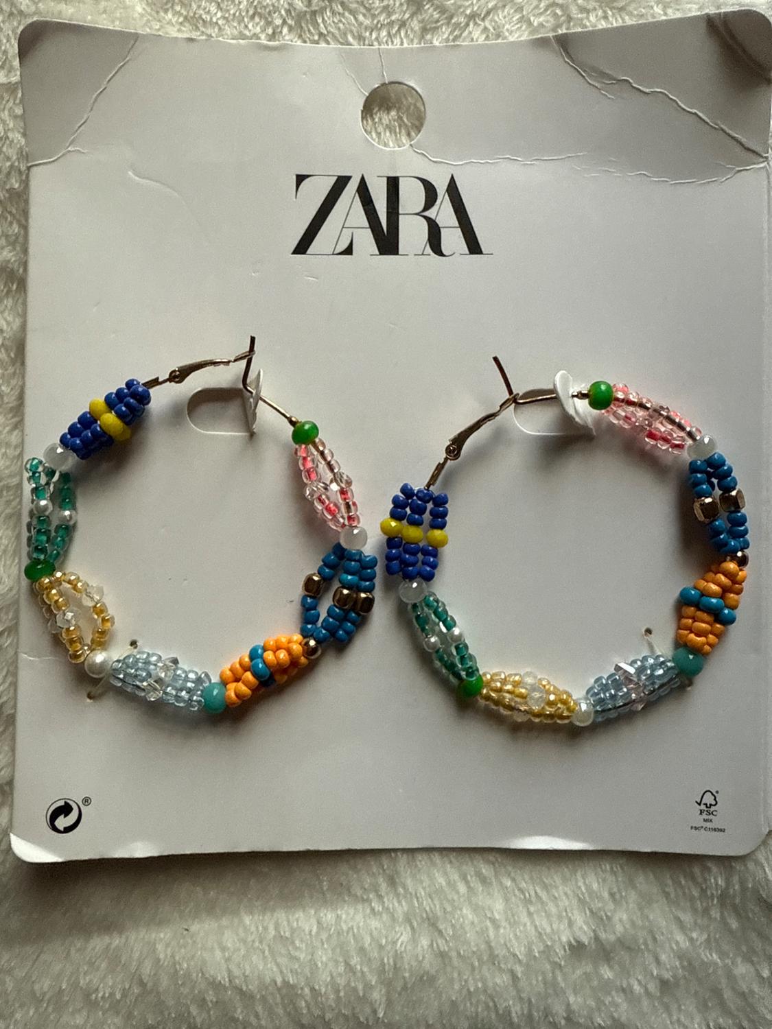 Hoop earrings with colorful beads on a white stand with the "ZARA" logo at the top. Soft fabric background.