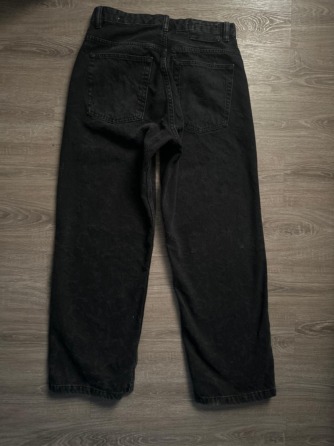 Black denim pants laid on a light wooden floor, viewed from the back, showing back pockets and belt loops.