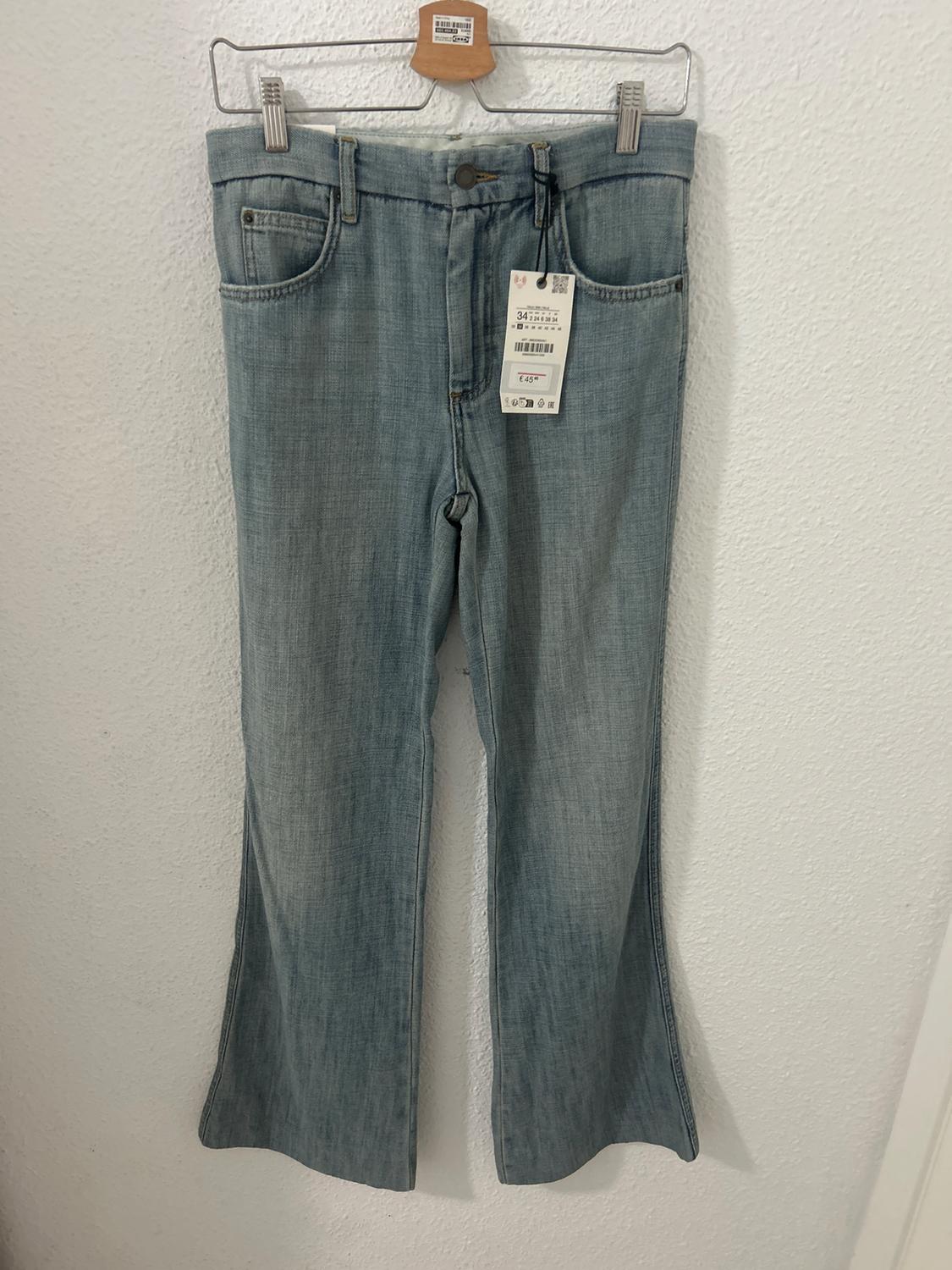 Blue denim jeans hanging on a wooden hanger, with a visible price tag. White wall background.