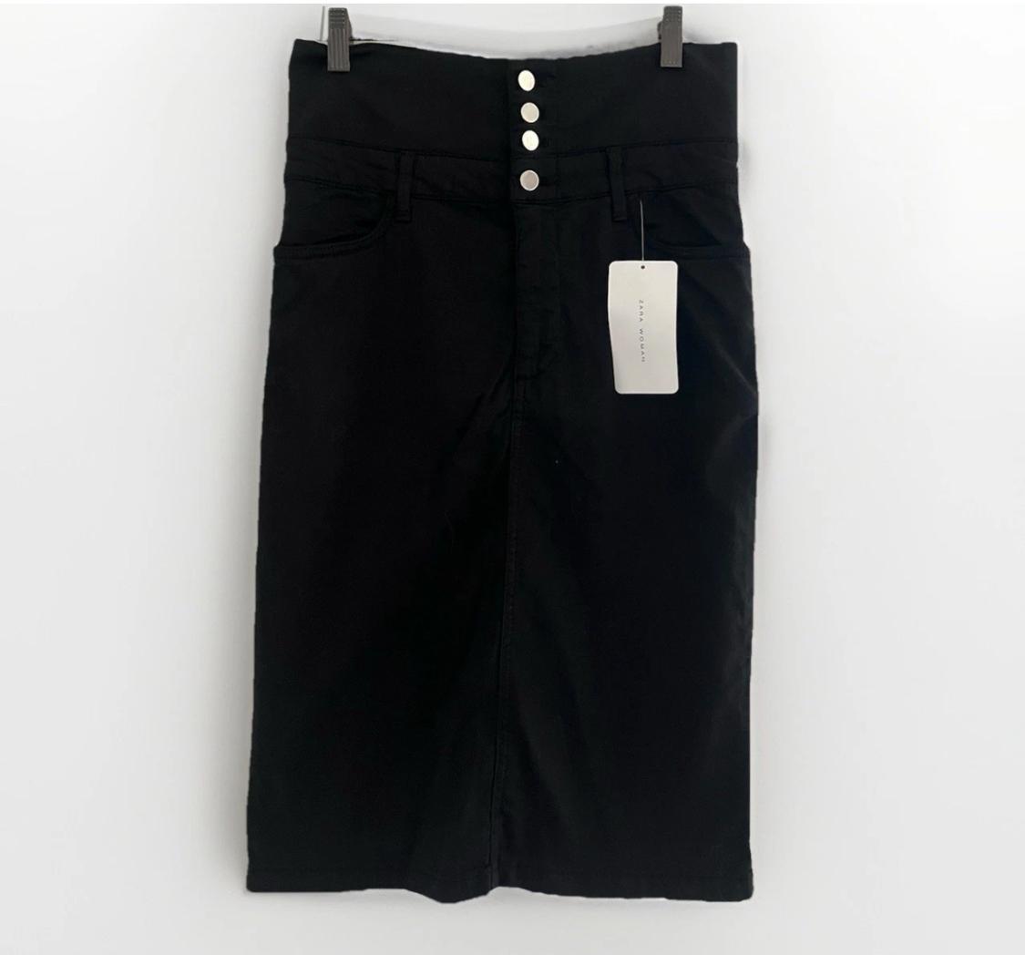 High-waisted black skirt with four front metal buttons, hanging on a hanger against a white background. Label with text ZARA WOMAN.