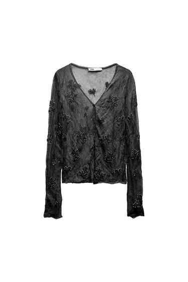 Sheer black blouse with floral embroidery and beads, long sleeves and V-neck, on a white background. Visible tag on the neck.