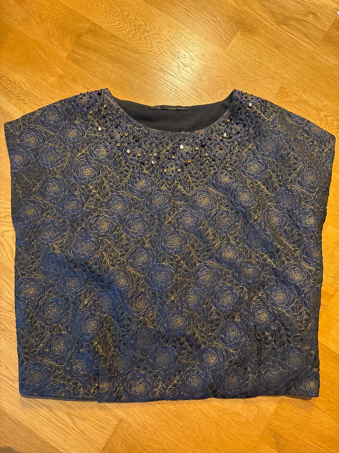 Blue blouse with gold floral embroidery and diamante details on the collar, on a light wood background.