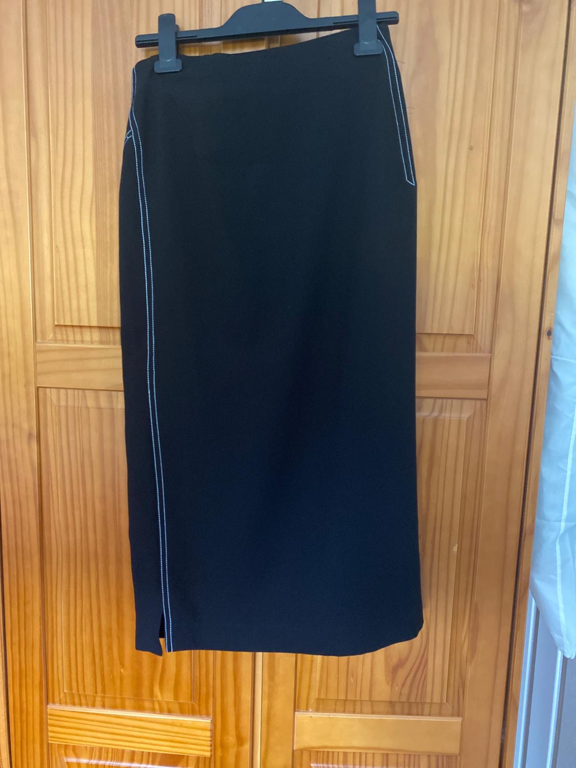 Long black skirt with white stitching on the side, hanging on a black hanger over a light wooden door.