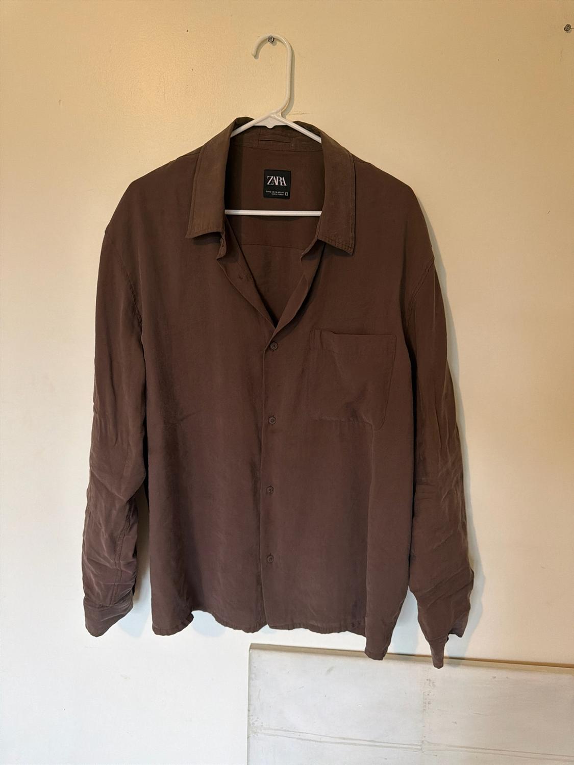 Long-sleeved brown button-up shirt with a front pocket, hanging on a white hanger against a light-coloured wall. Label with the text ZARA.