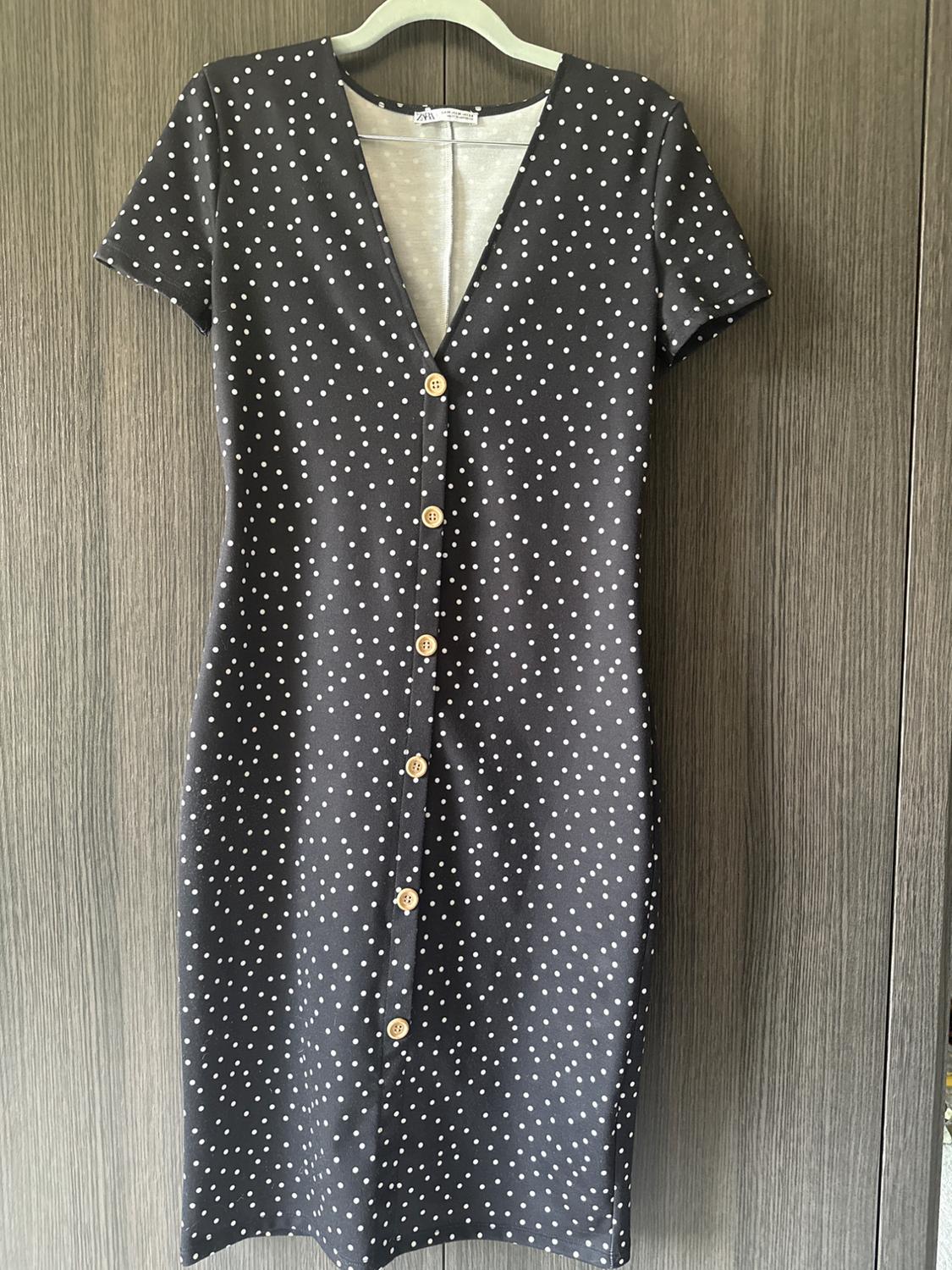 Casual black ZARA dress with white polka dots, short sleeves and front buttons, hanging on a hanger against a wooden background.