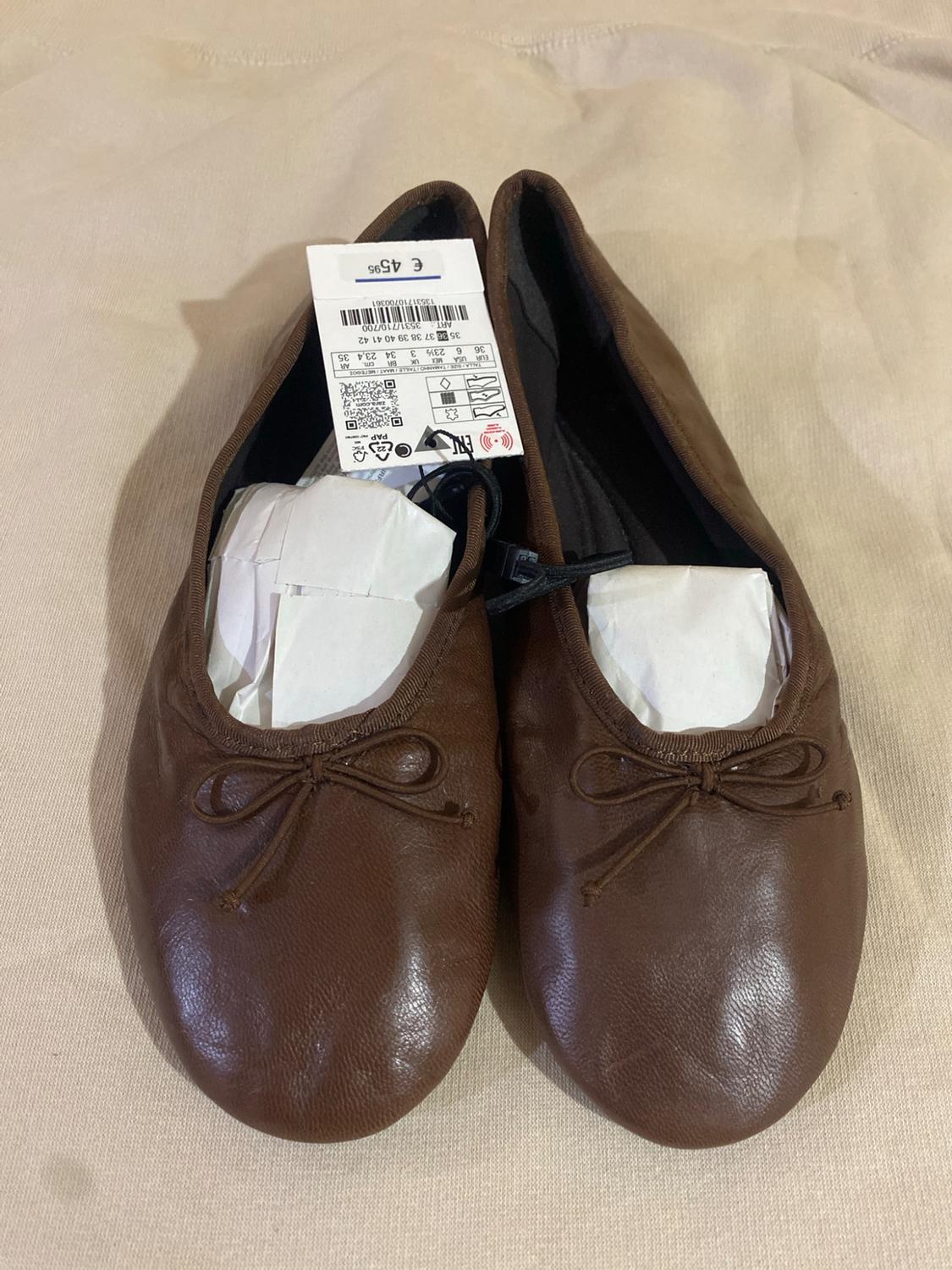 Pair of brown ballet flats with a bow on beige fabric. They have paper inside and a tag with the text: €45.95.