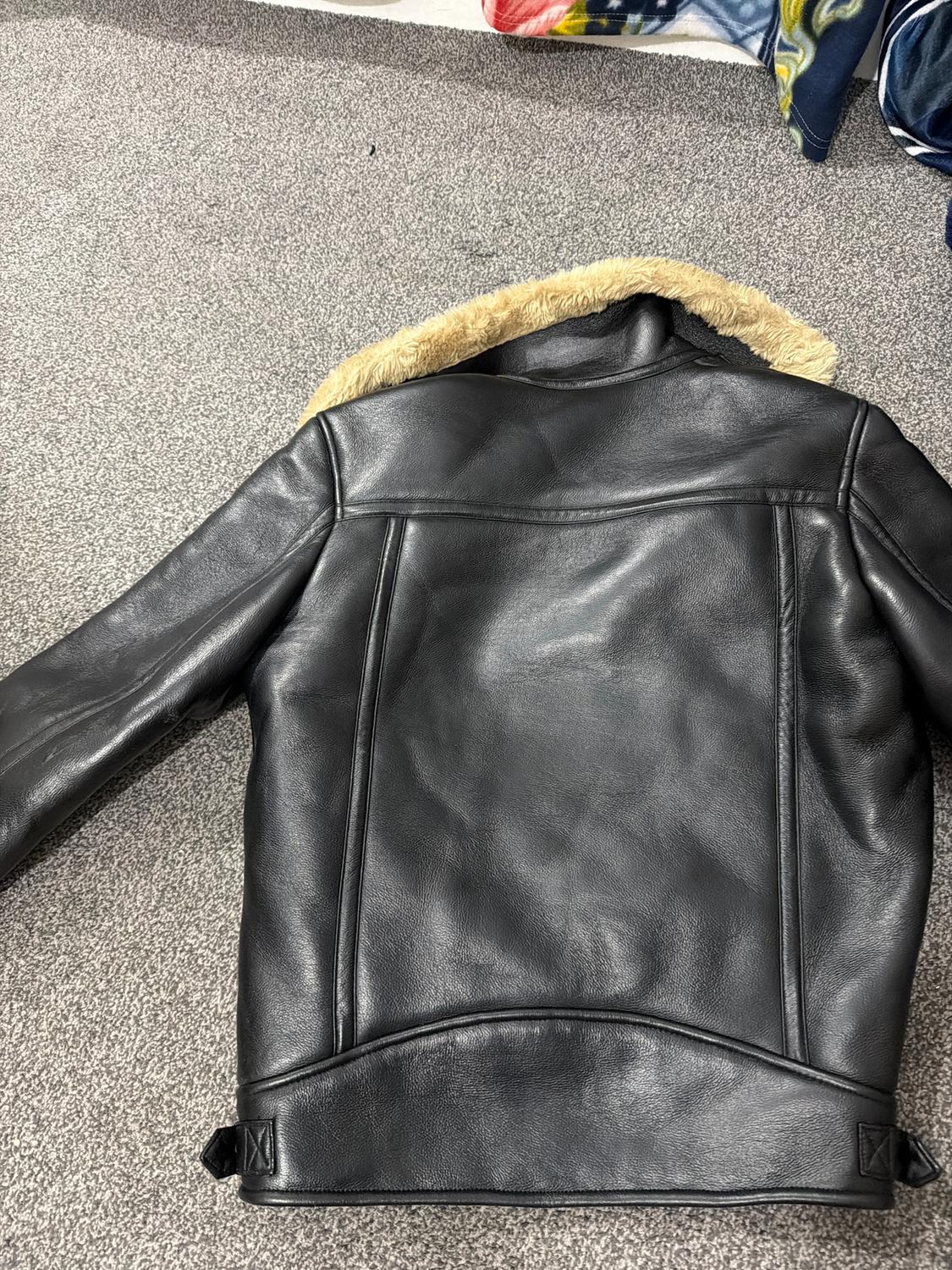 Black leather jacket with a light fur collar, viewed from behind on a grey rug. Part of a colourful blanket visible in the background.