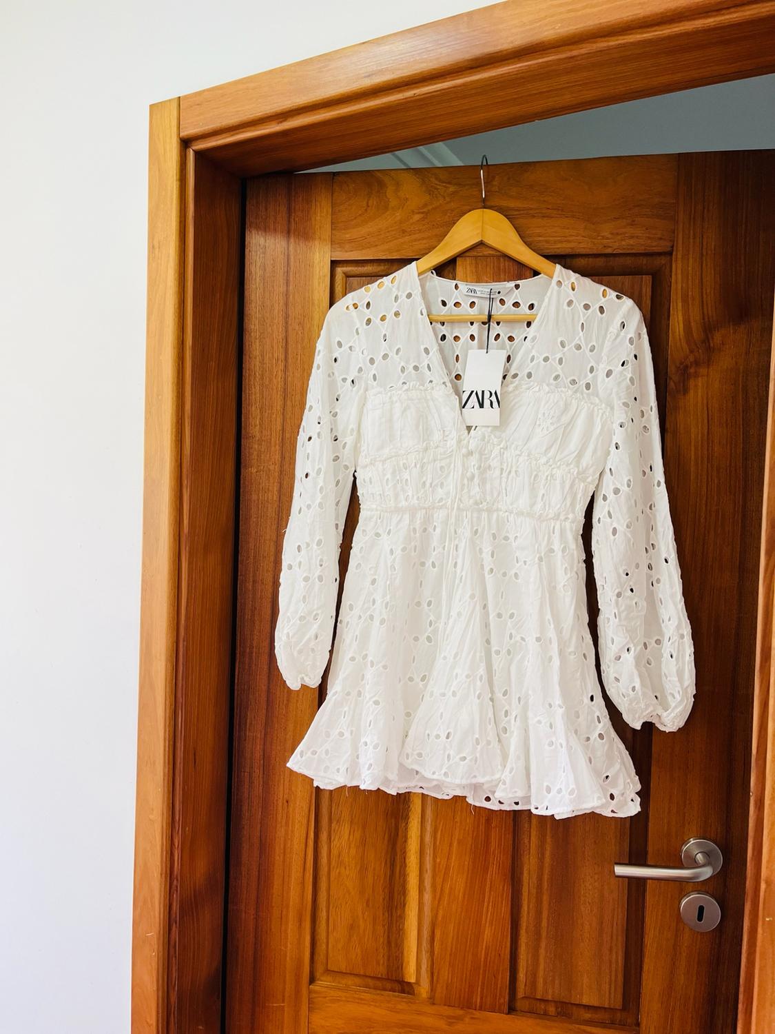 White long-sleeved short dress with openwork embroidery, hanging on a wooden door. Label visible with the text ZARA.