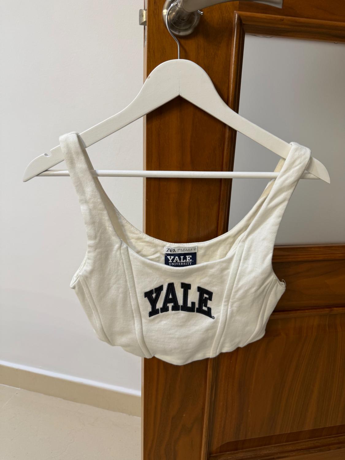 Άσπρο τοπ με τη λέξη "YALE" σε μαύρα γράμματα κρεμασμένο σε μια άσπρη κρεμάστρα πάνω από μια ξύλινη πόρτα.