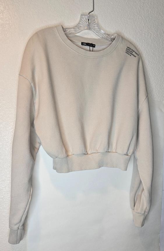 Beige sweatshirt hanging on a clear hanger. Text on the shoulder: "LIMITED SPORT BASICS, FUTURE MOVEMENT, LIMITED EDITION, LOOKING AT".