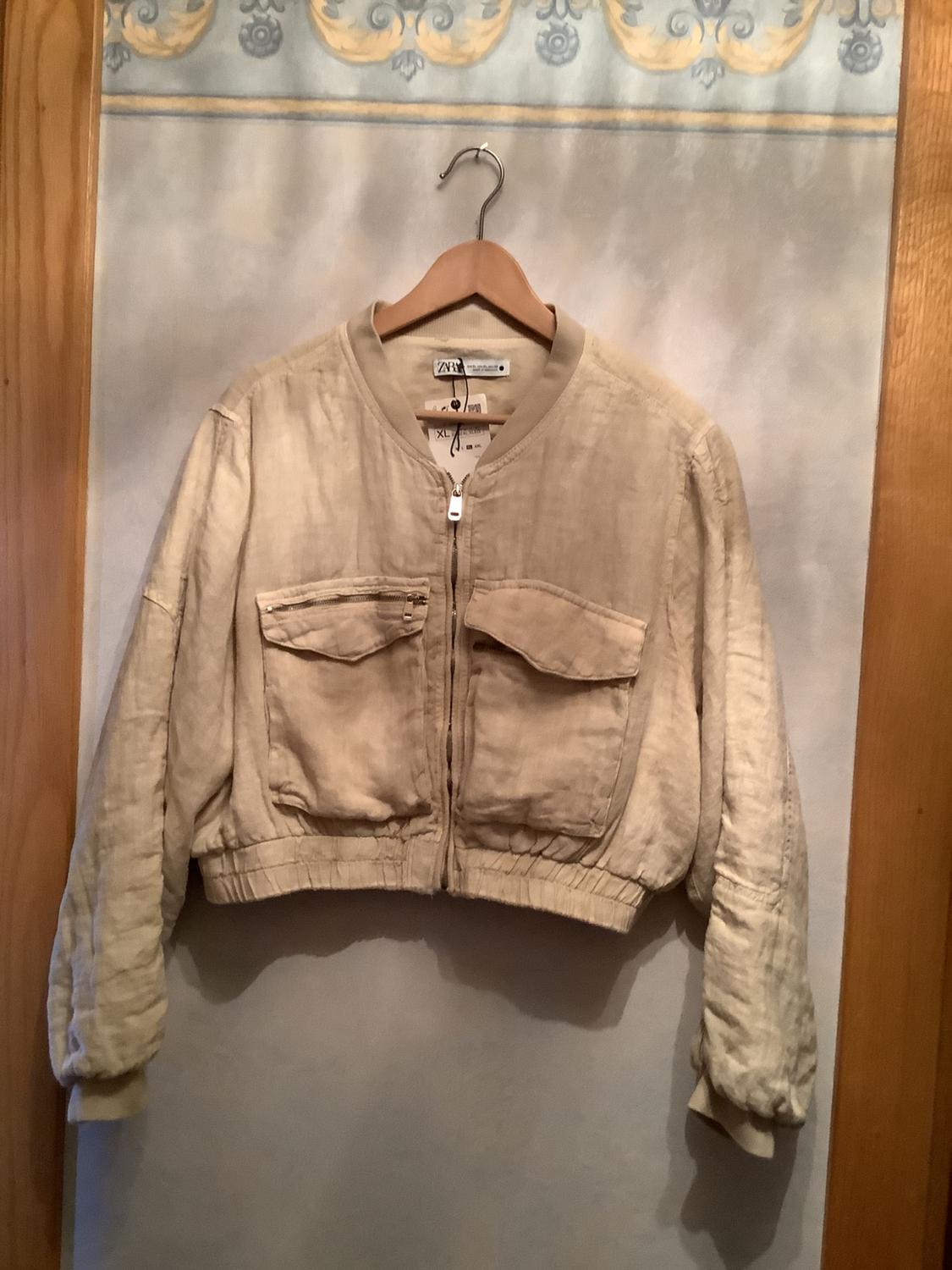 Beige linen jacket with a zip and two front pockets, hanging on a wooden hanger against a decorative background.