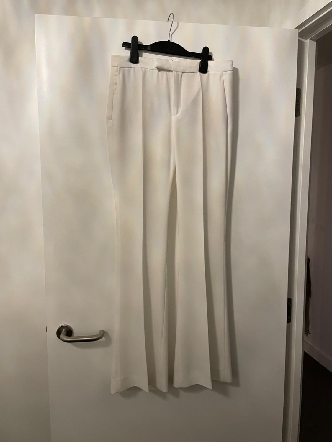 White tailored trousers with a crease, hanging on a black hanger over a white door.