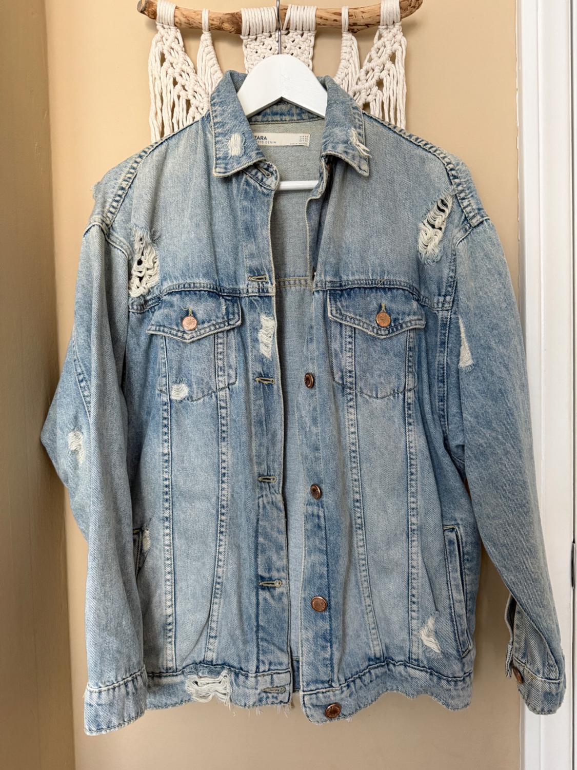 Worn blue denim jacket hanging on a white hanger against a beige background with macrame details.