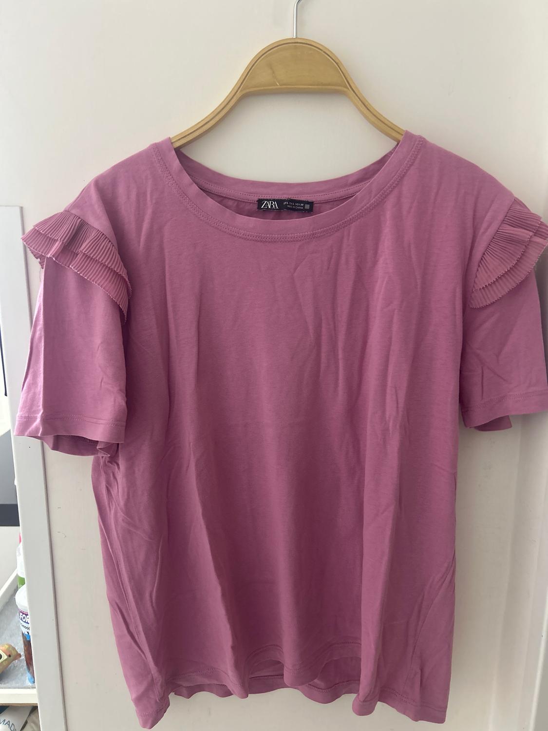 Pink T-shirt with ruffled sleeves, hanging on a wooden hanger over a white door. Label visible on the collar.