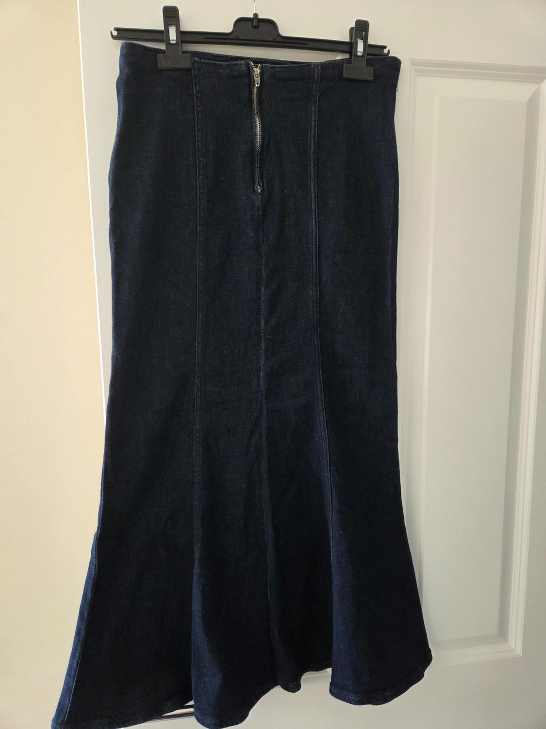 Dark blue denim long skirt with vertical seams and a front zip, hanging on a black hanger over a white door.