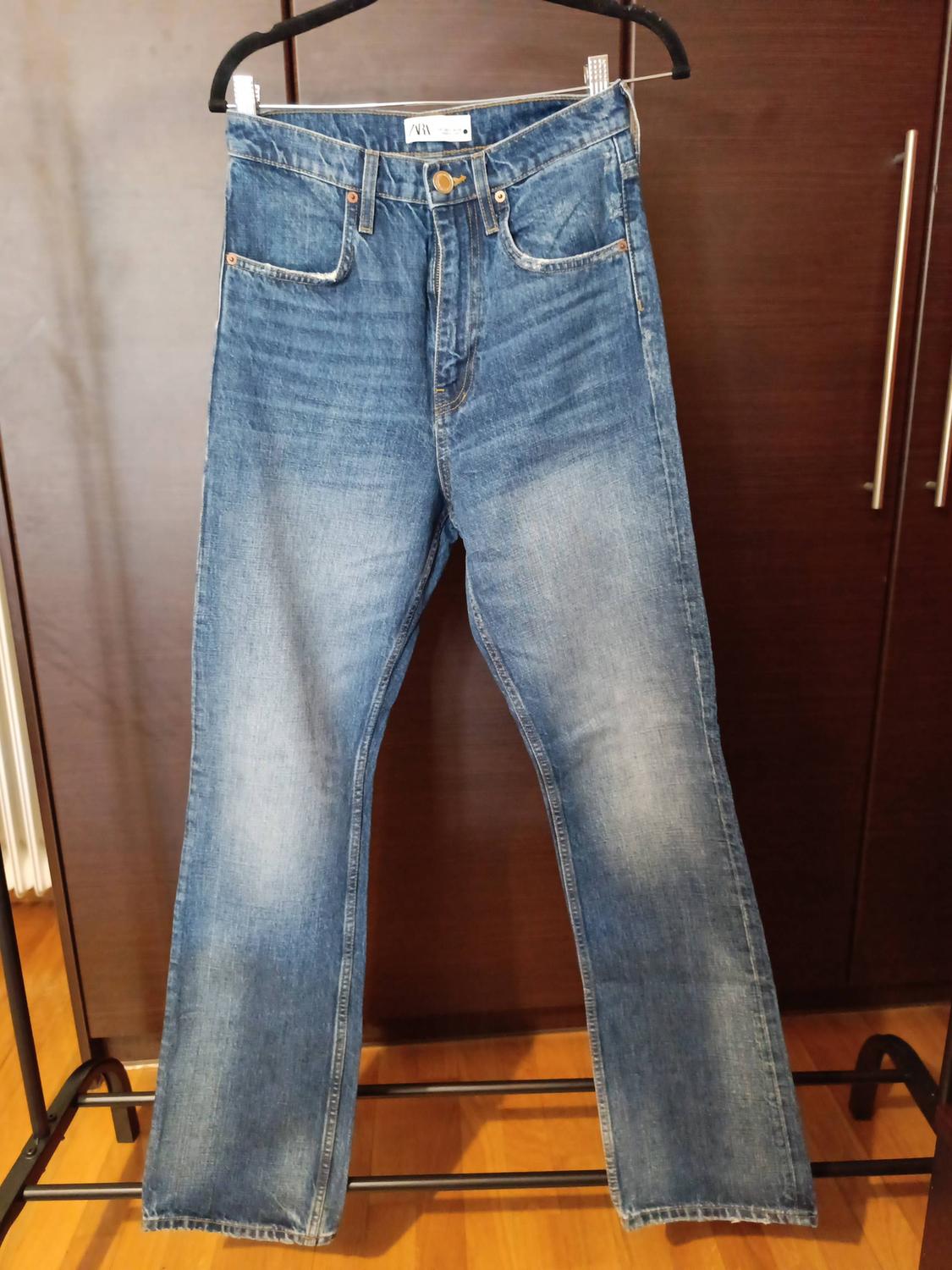 Blue jeans hanging on a black hanger against a dark wardrobe background. Natural light illuminates the wooden floor.