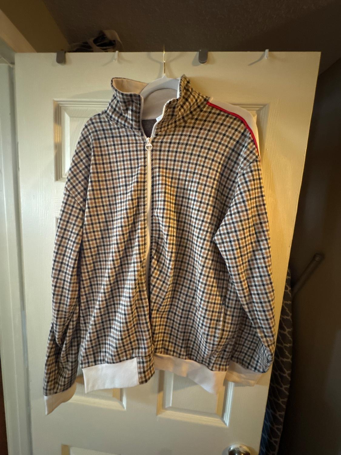 Brown and white checked jacket with a zip and high collar, hanging on a white door. Red detail on the shoulder.