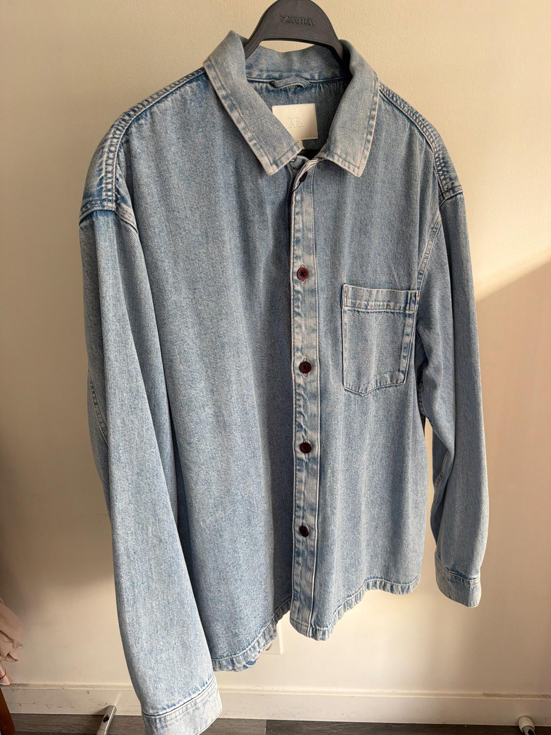 Light blue denim shirt hanging on a black hanger with visible branding. Beige wall background and wooden floor.