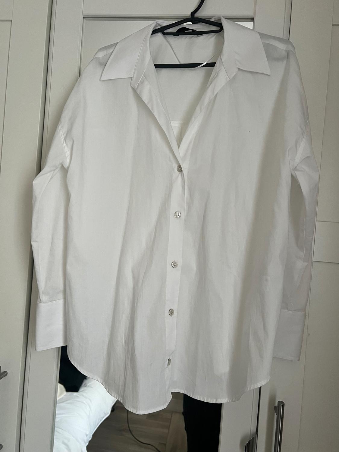 White button-down shirt hanging on a black hanger in front of a white wardrobe. Background with partially visible bed.