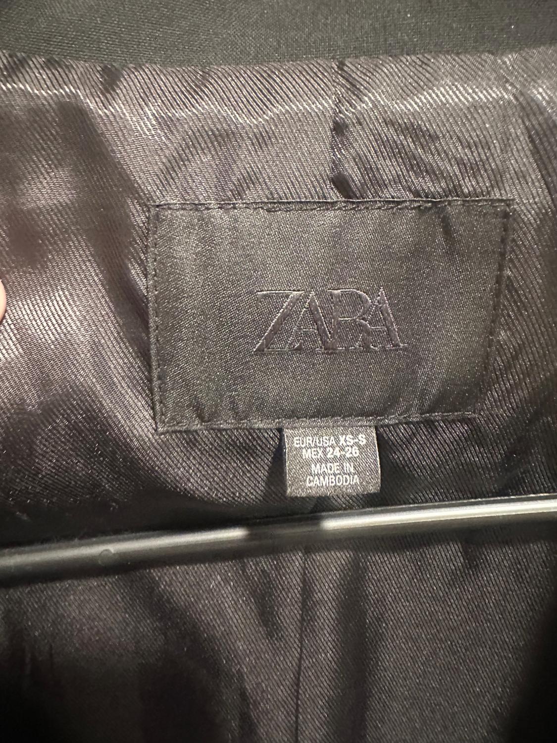 Black label on garment lining with the text ZARA EUR/USA XS-S MEX 24-26 MADE IN CAMBODIA, with a visible hanger.