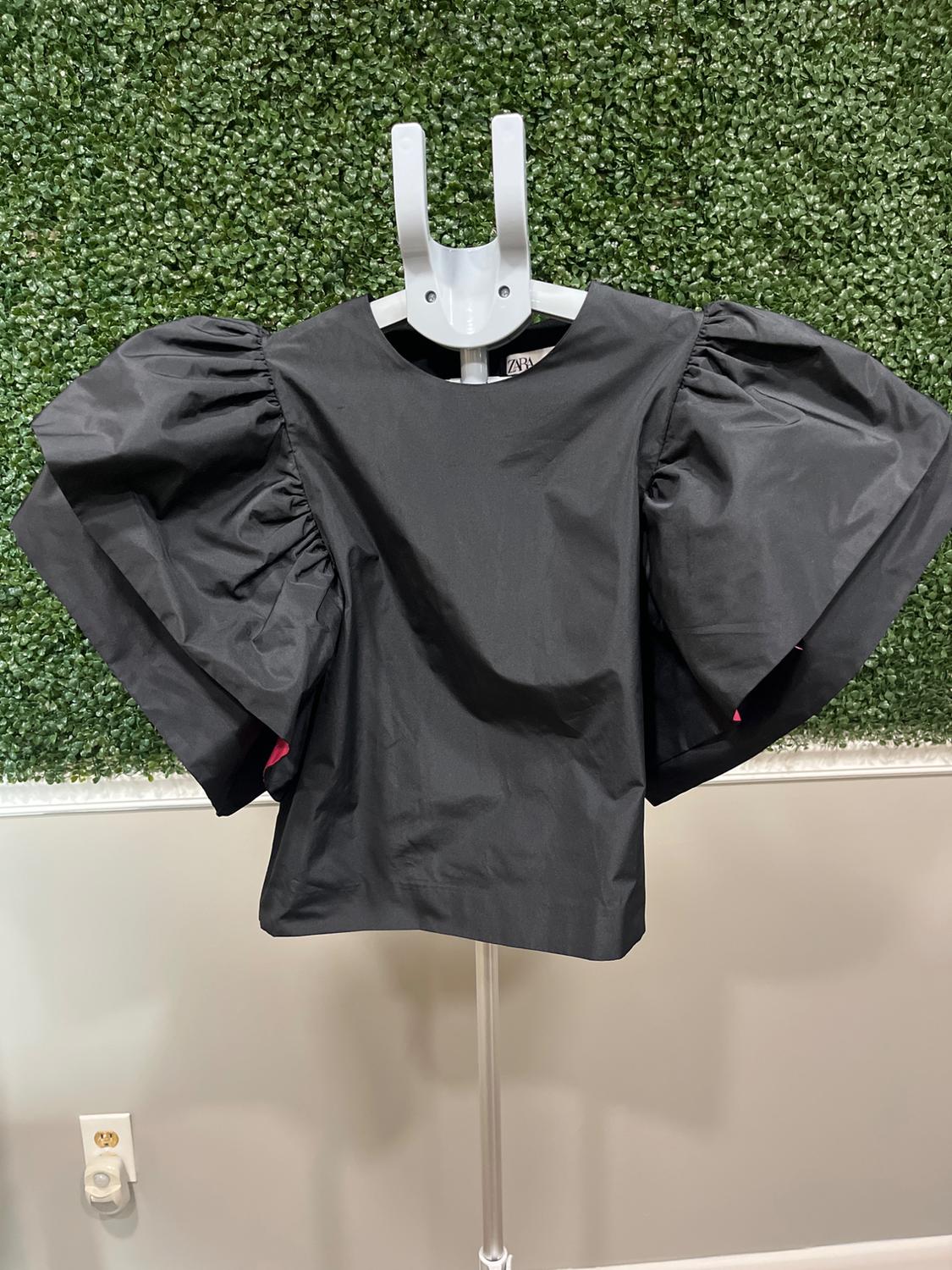 Black blouse with puff sleeves on a mannequin, green leaf wall background. Visible tag: "ZARA". Plug on the wall.