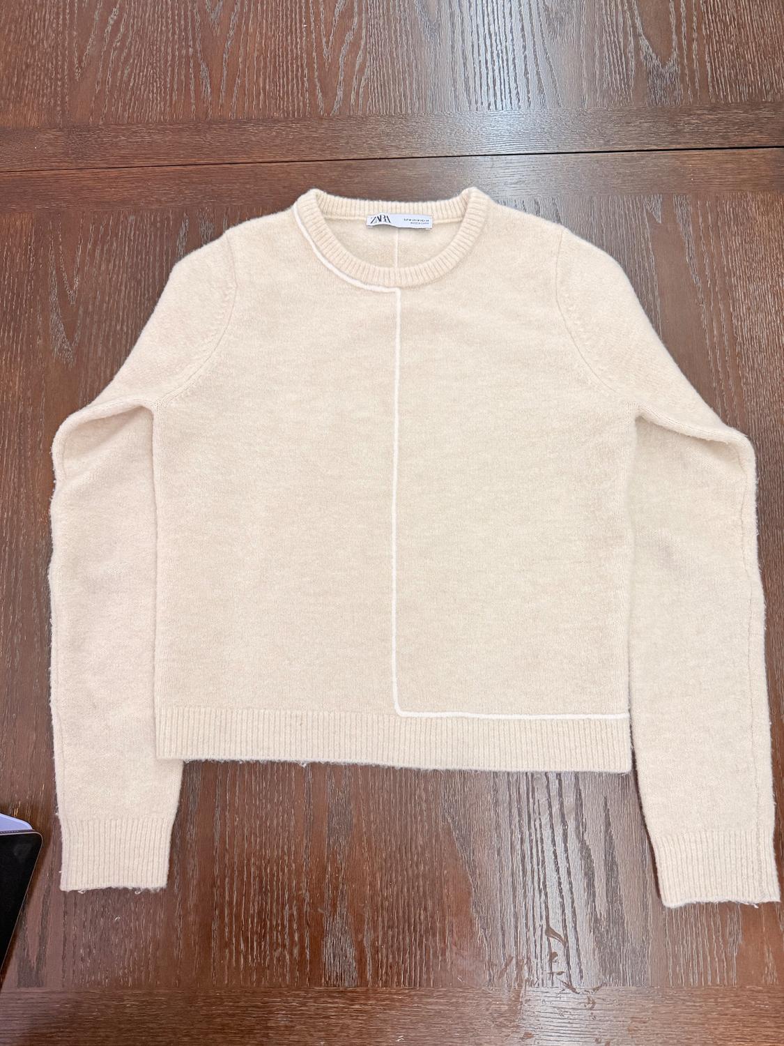 Long-sleeved beige sweater on dark wooden table, with visible "ZARA" tag on the collar.