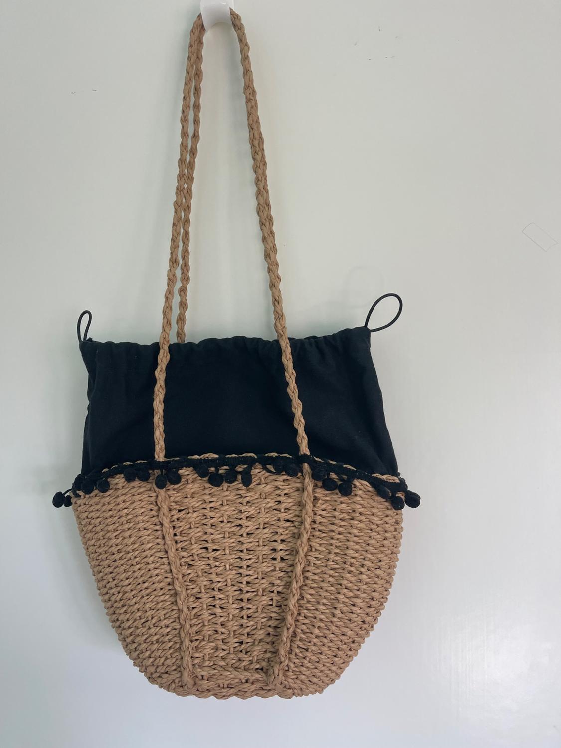 Woven straw bag with long handles and black fabric top with tassel details, hanging against a white background.