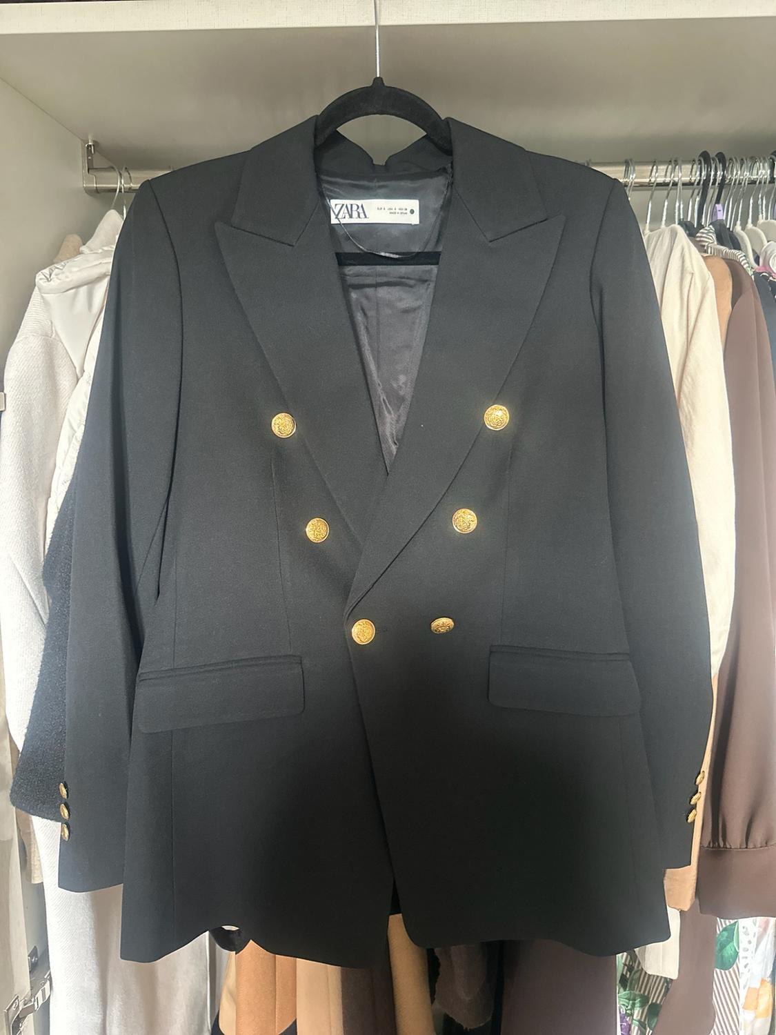 Black double-breasted blazer with gold buttons hanging in a wardrobe with other clothes in the background. Label with the text ZARA.
