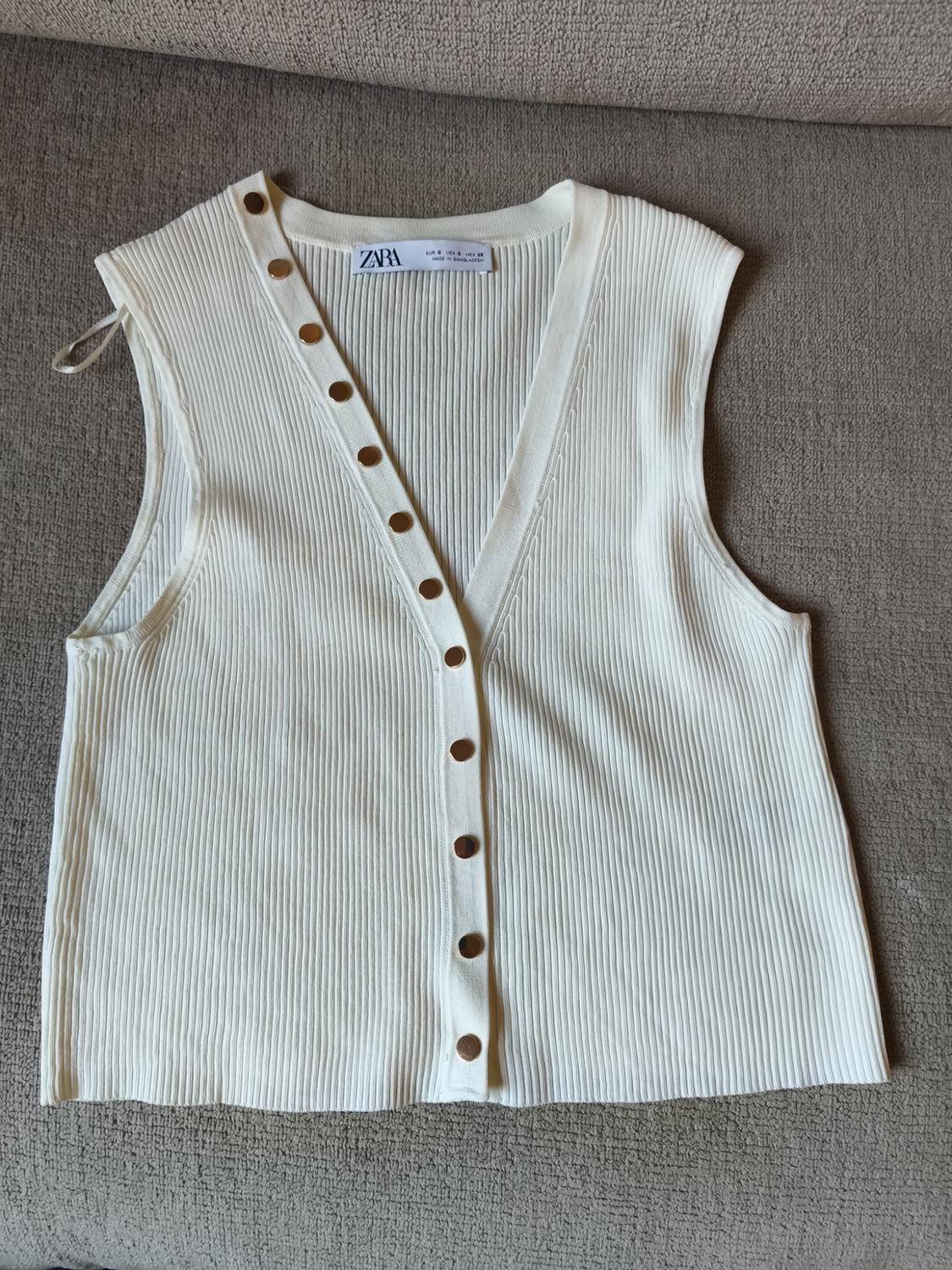 White knitted waistcoat with brown buttons on a grey sofa. Visible label with the text "ZARA".