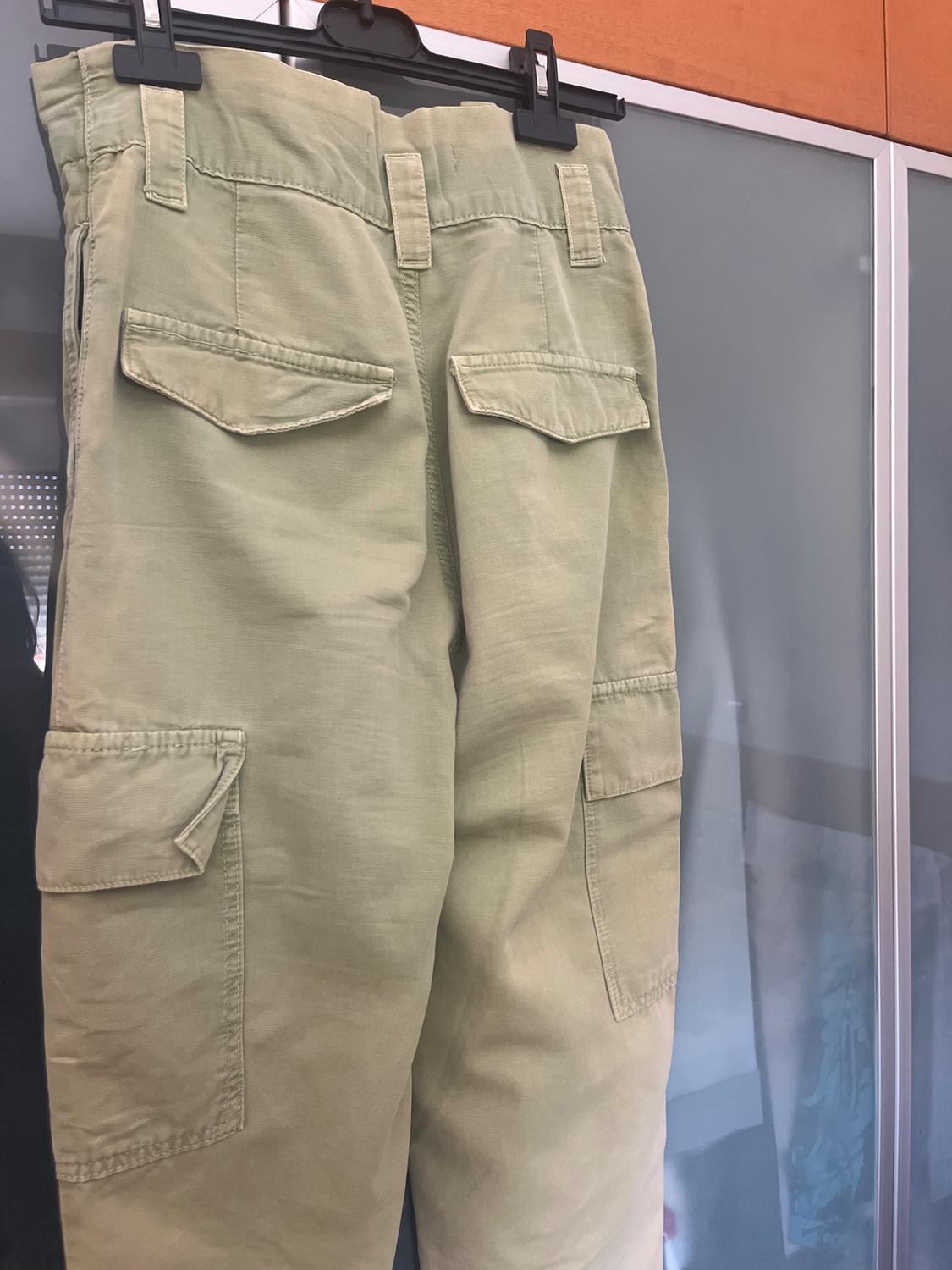 Green cargo trousers hanging on a black hanger in front of a glass panel. Rear view with flap pockets.