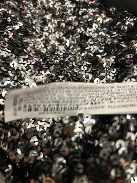 Fabric with silver and black sequins in the foreground, label with blurred text in various languages on the fabric.