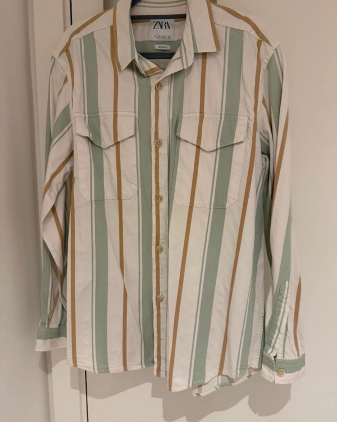 Long-sleeved shirt with vertical green, mustard and white stripes, hanging. Two front pockets. Label with the text ZARA.