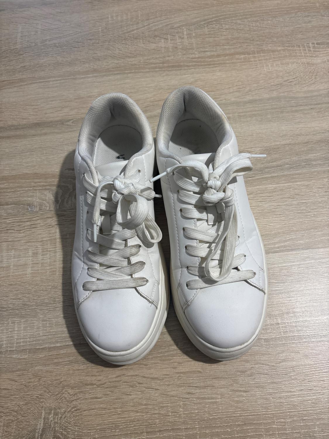 White lace-up trainers on a light wooden floor. Top view, showing stitching details and a simple design.