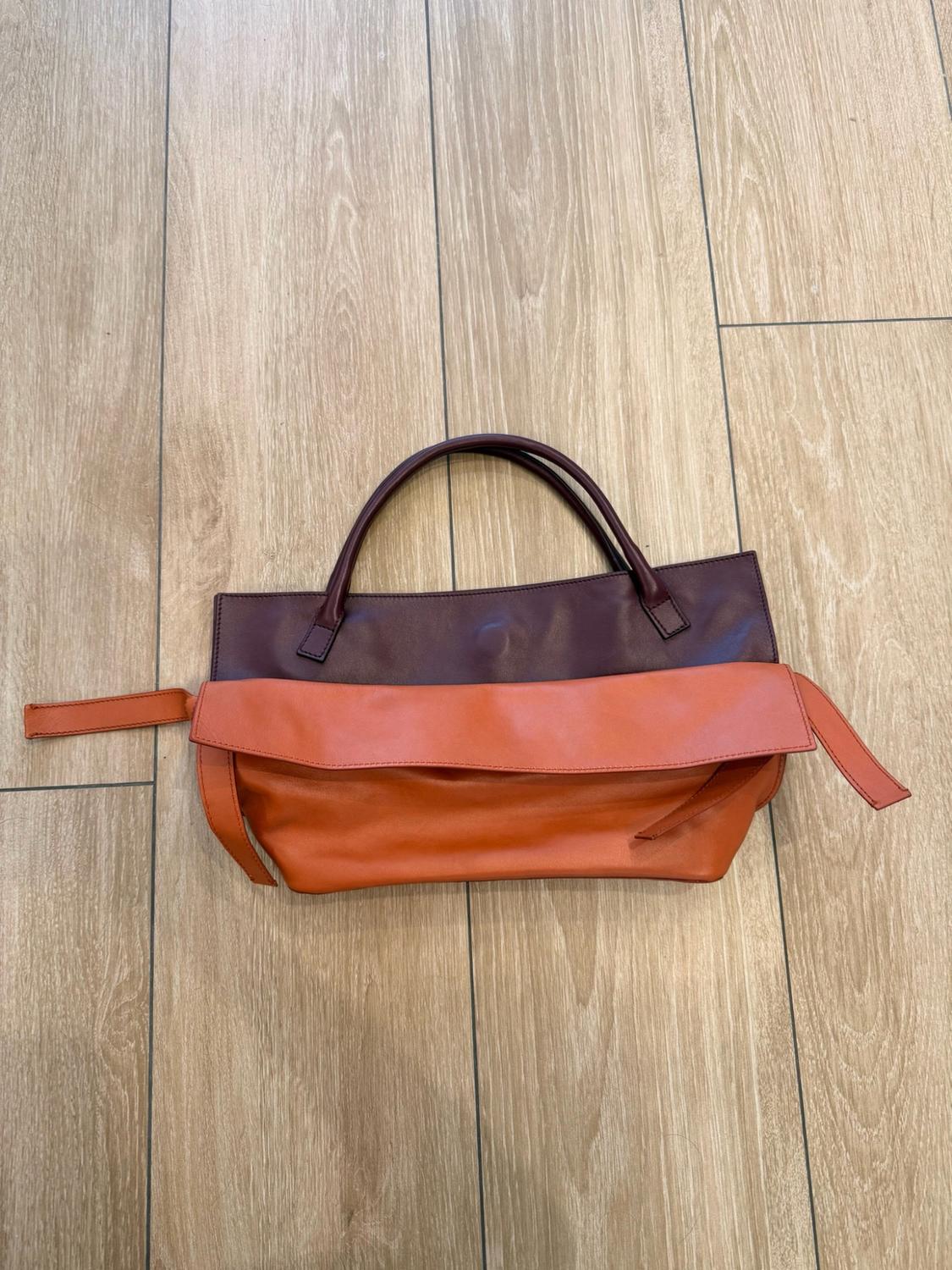 Handbag in burgundy and orange tones with short handles, resting on a light wood floor in a casual style.