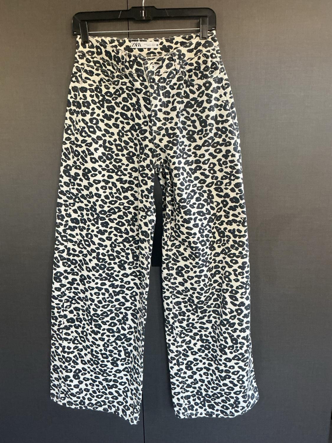 Black and white animal print jeans hanging on a black hanger against a dark background. Tag text: Zara.