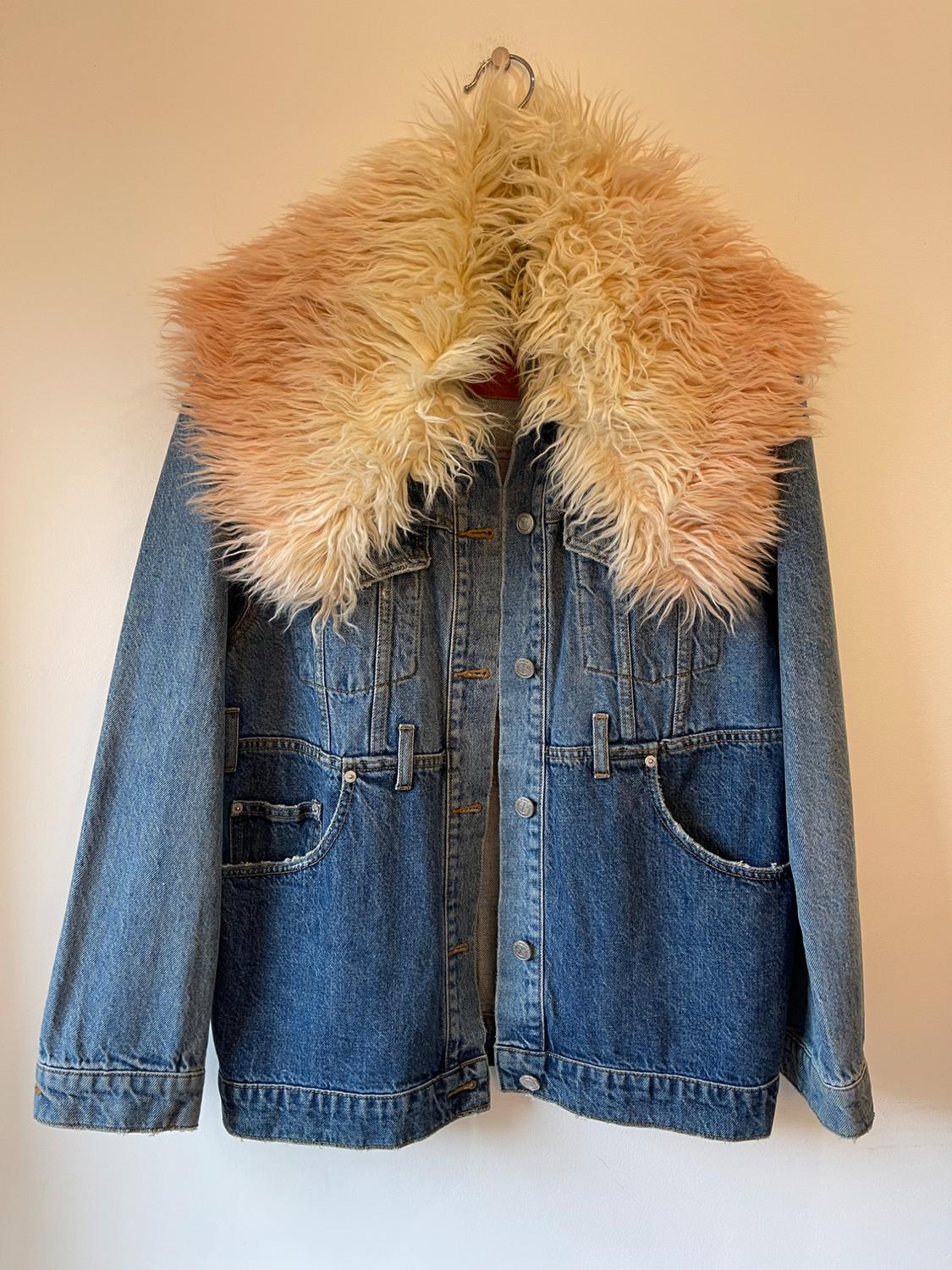 Blue denim jacket with a faux fur collar in beige and pink tones, hanging on a hanger against a white background.
