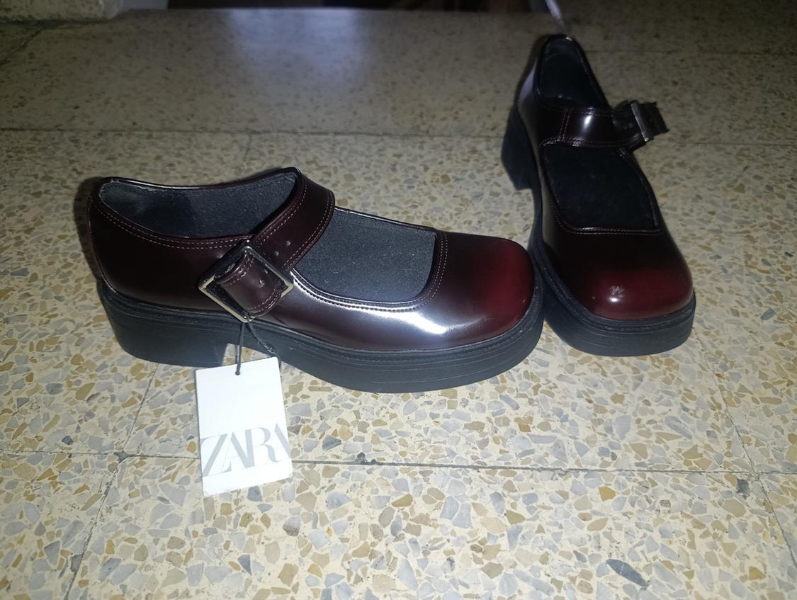 Shiny burgundy Mary Jane shoes with a black platform and buckle, on a terrazzo floor. Includes a label with the text ZARA.