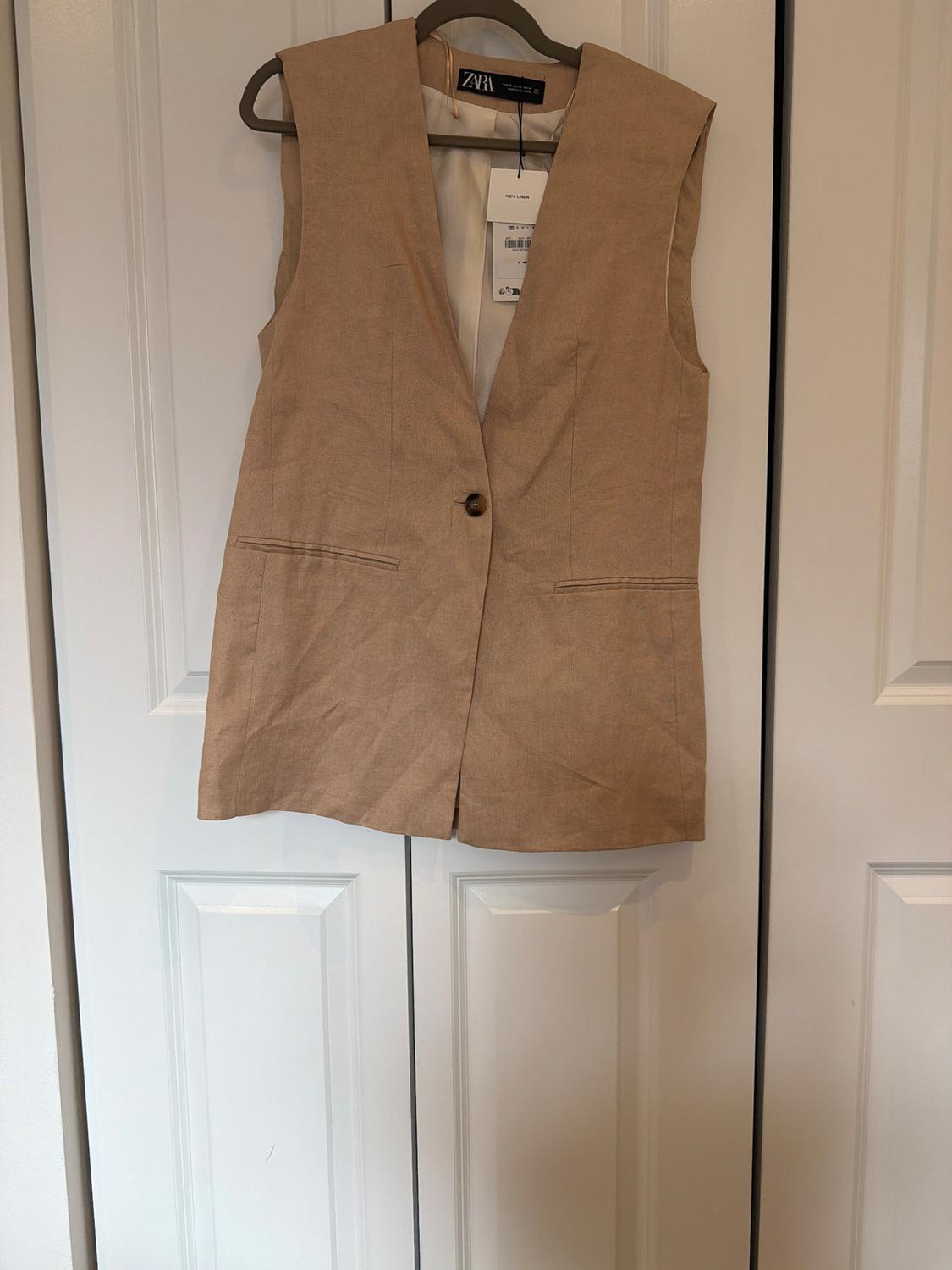 Beige one-button vest hanging on a hanger on a white door. Visible tags with the text ZARA and 100% LINEN.