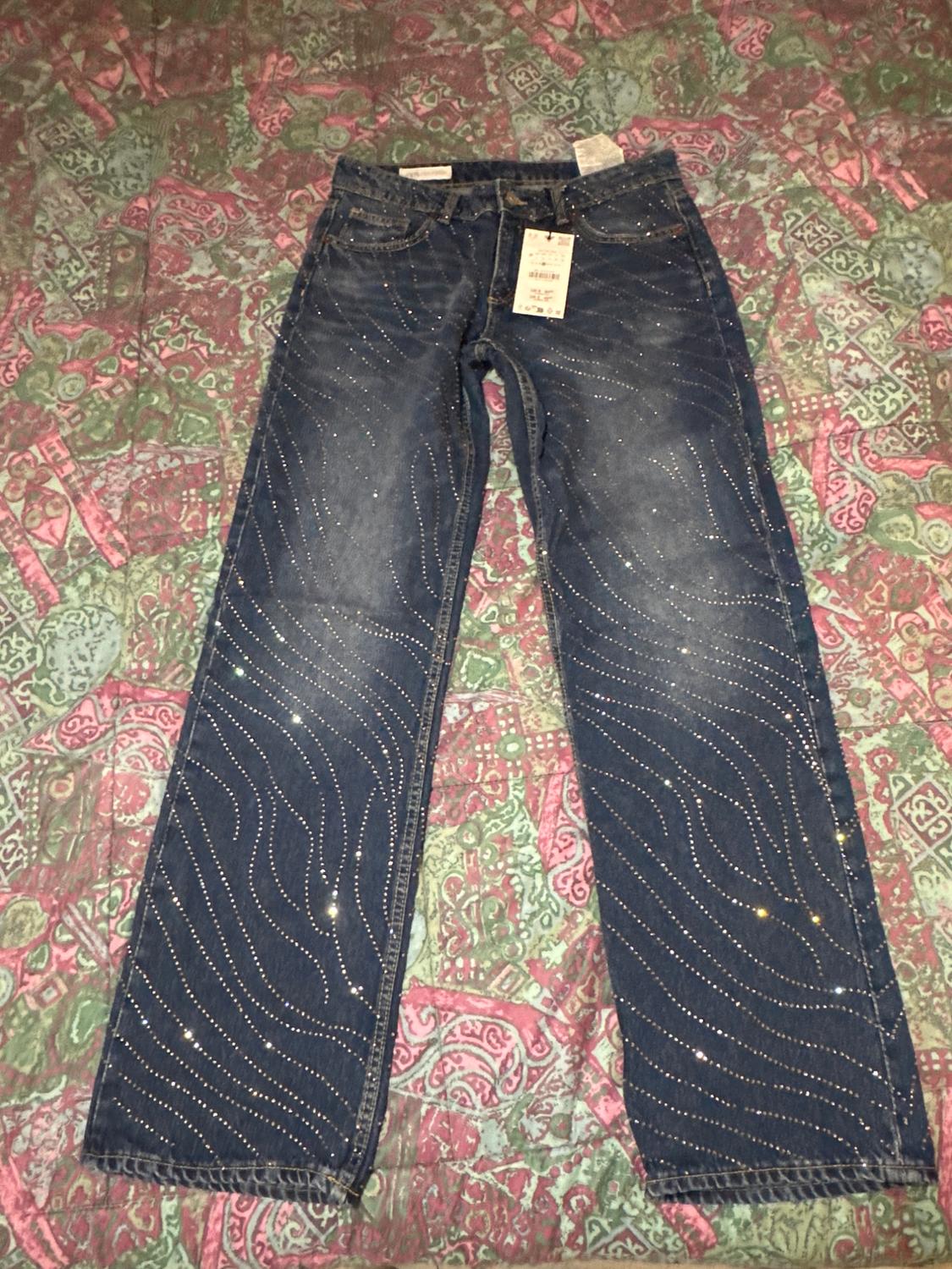 Dark blue jeans with a shimmering wave design on a colourful quilt. Hang tag visible at the top.