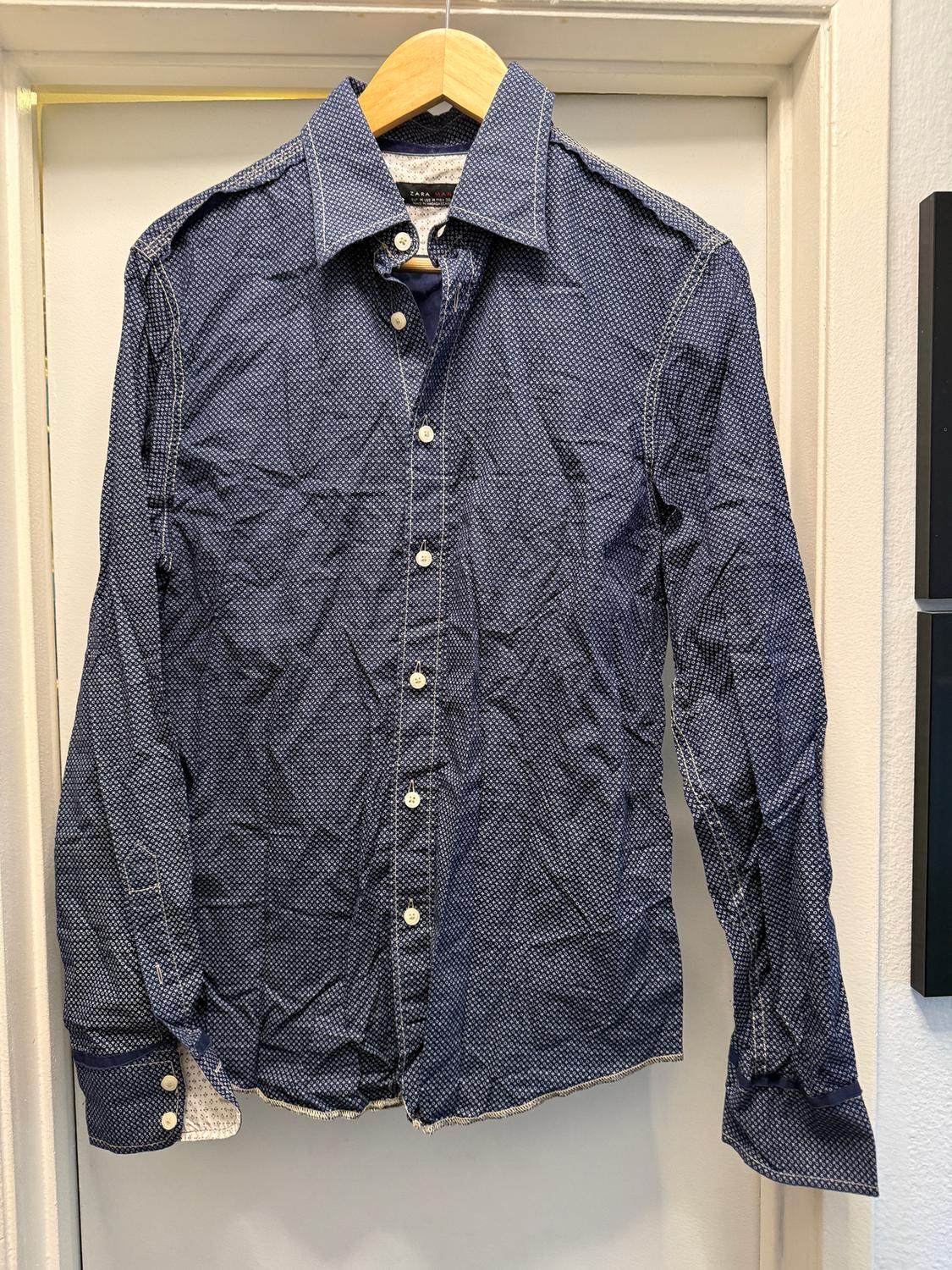 Blue shirt with a white polka dot pattern, hanging on a wooden hanger. White buttons and visible stitching.