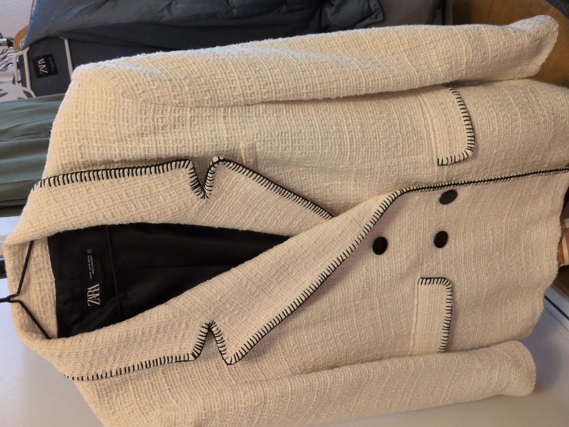 Beige textured fabric coat with black buttons and dark trim, hanging on a hanger. Visible label: ZARA.