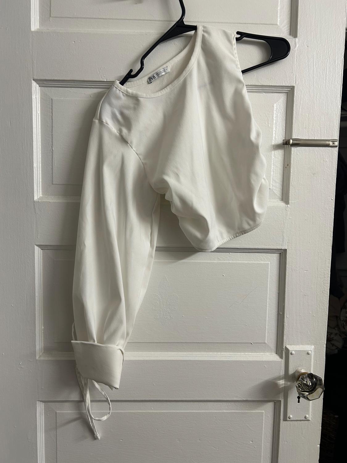 Asymmetrical white top with one long sleeve and a tie at the cuff, hanging from a black hanger on a white door. ZARA label.