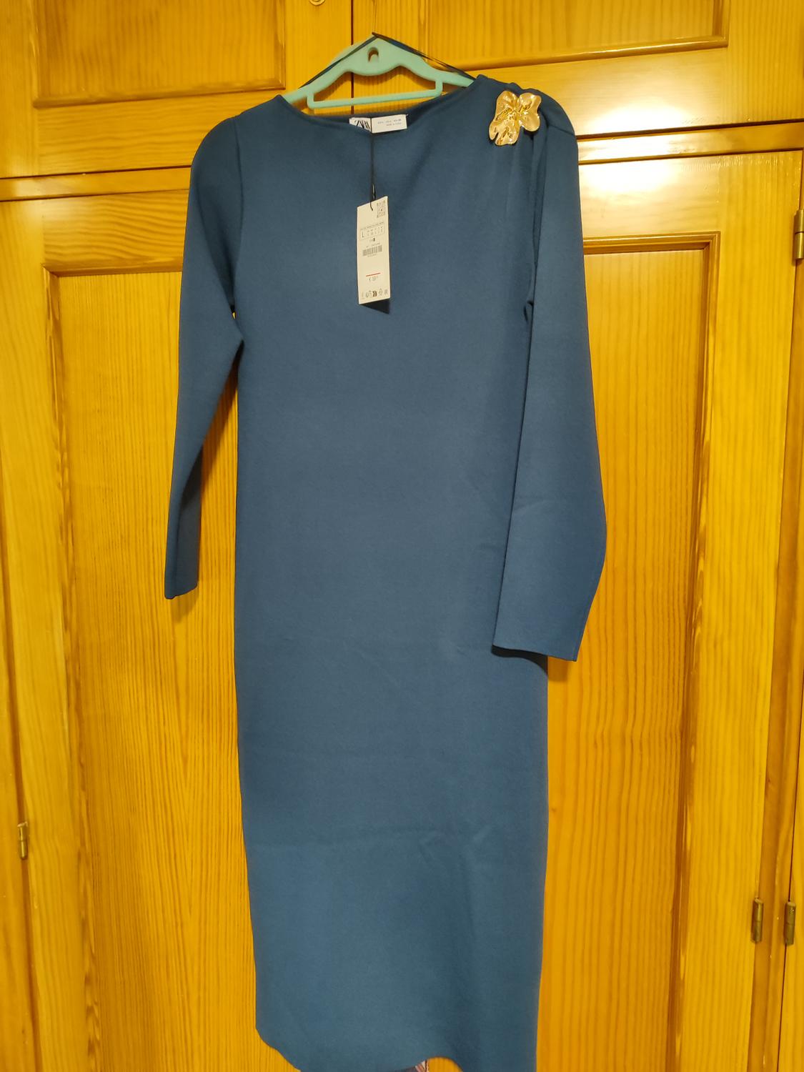 Long-sleeved blue dress hanging on a hanger, with a gold floral detail on the shoulder and a visible tag, against a light wooden background.